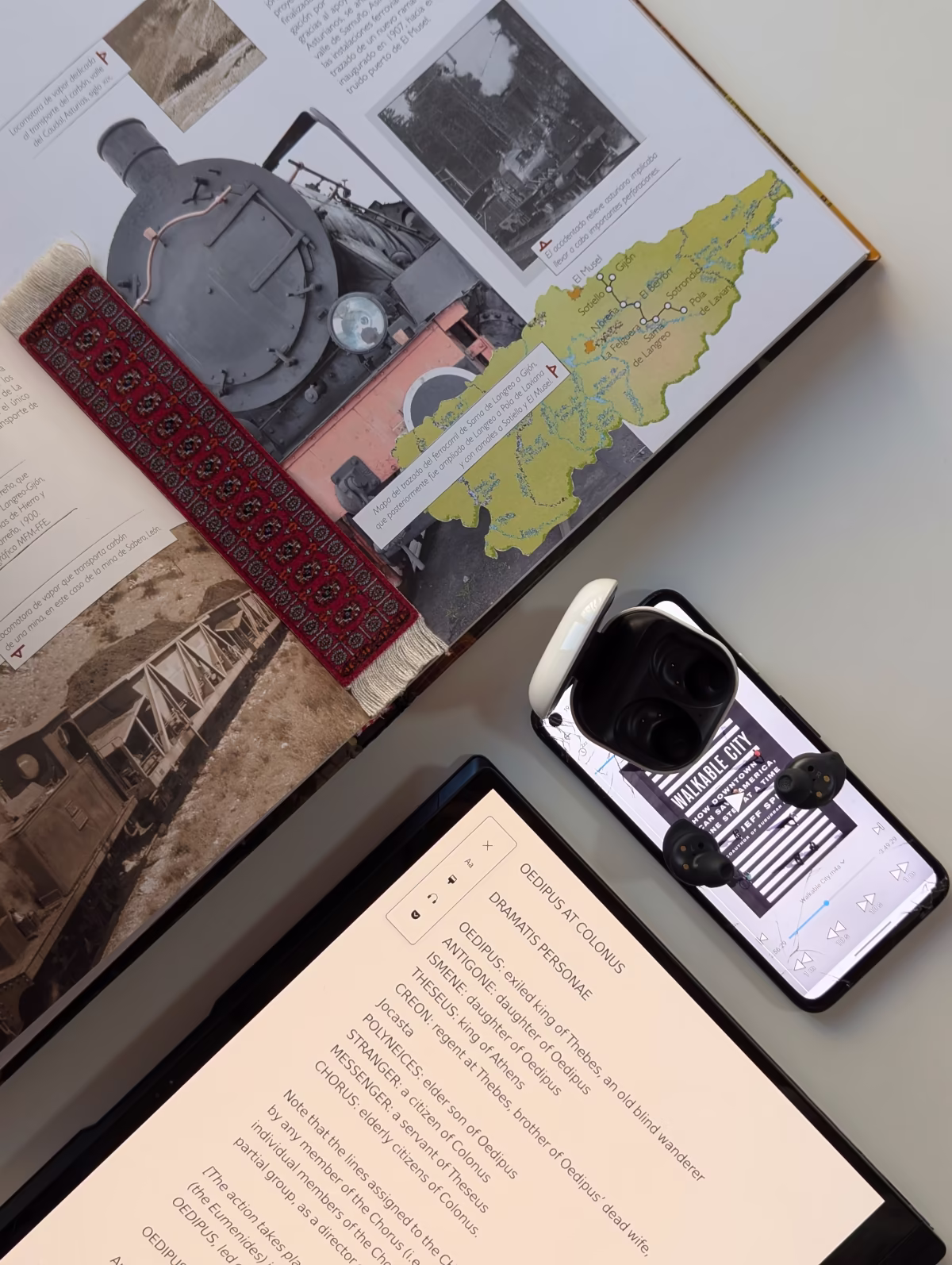 Una vista áerea de un libro, un portátil-tableta, y un móvil con auriculares sobre una mesa. El libro físico, Historia del Ferrocarril de España, es de tapa dura con ilustraciones de varios trenes del siglo 19 y mapas relacionados con cajas de texto, y tiene un marcador de libros. El portátil está plegado como una tableta y muestra Oedipus At Colonus, en el modo lector de Firefox, con opciones para cambiar el texto y el diseño, y usar la lectora de voz. El móvil tiene abierto el audiolibro Walkable City, con auriculares y su estuche puesto sobre el móvil.